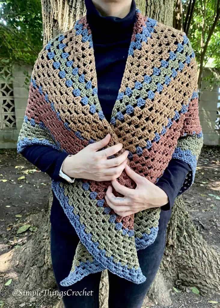 Person wearing the crochet Autumn Woods Wrap shawl in blue, brown and cream granny stitch outdoors in the woods